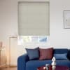 Beige cellular blind fully lowered over window, textured ribs; filtering daylight and covering most glass; modern living room with blue sofa, cushions, floor lamp, and a rooftop view outside.