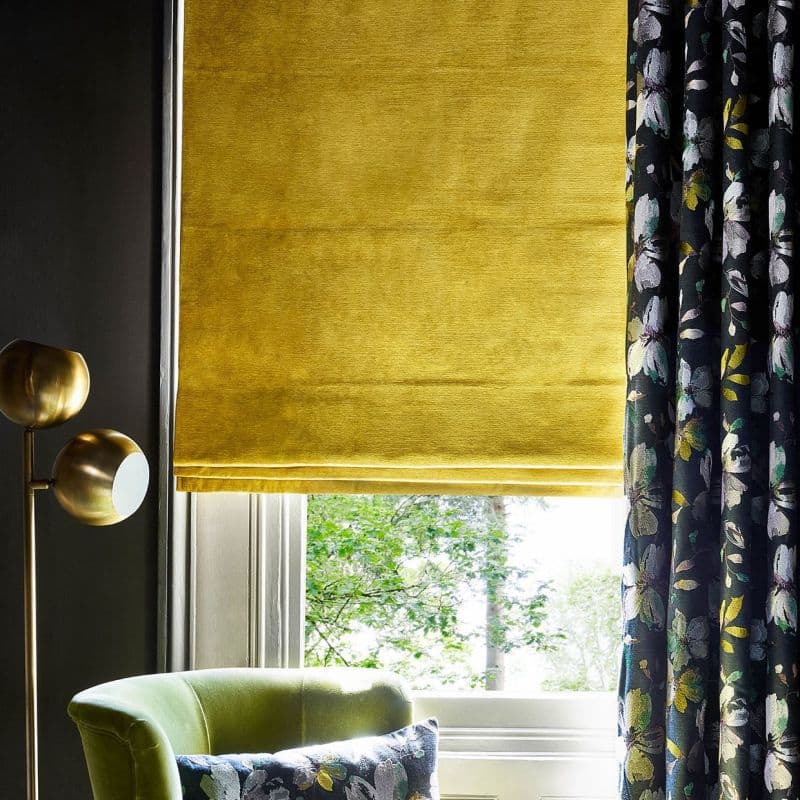 Yellow velvet Roman blind lowered, paired with a dark floral curtain drawn right; filtering daylight in a living-room corner with green armchair, brass lamp and a leafy outside view.