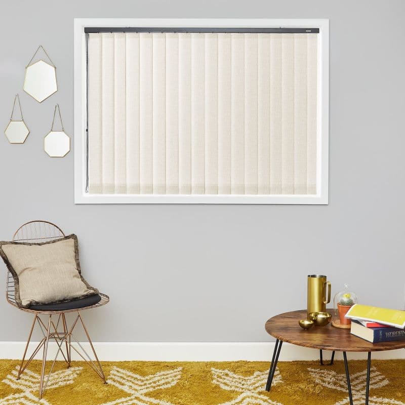 Beige vertical blinds, closed and diffusing light; fill a white-framed window on a grey wall, above a mustard rug beside a wire chair and small wooden table with books.