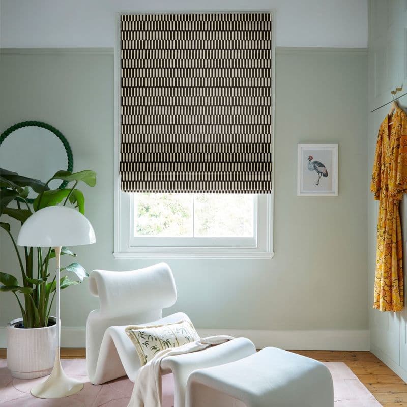 Patterned black-and-cream Roman blind lowered over a central sash window, filtering daylight; pale-green reading nook with sculptural white lounge chair, floor lamp, potted plant, framed bird print, yellow robe.