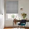 Grey Roman blind, lowered in soft folds, partially filtering daylight through the window; above a small desk in a bright home office with a teal chair and a vase of flowers.