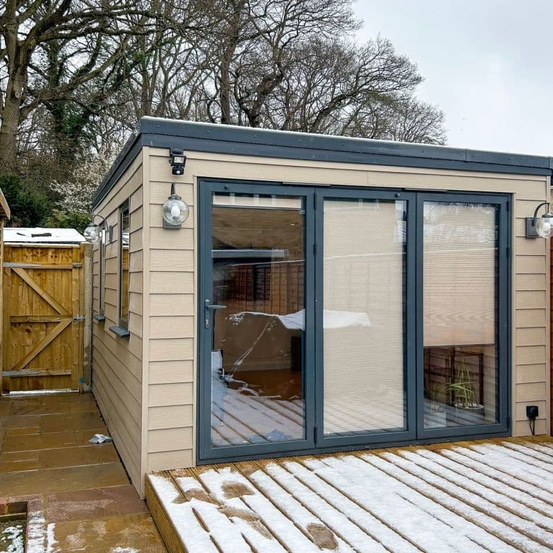 Beige pleated blinds, mounted inside glass double-door and adjacent pane, fully lowered and blocking view; on a grey-framed garden studio with beige cladding and a snowy wooden deck outside.