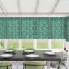 Dining table set with stacked white plates, green glass candlesticks and small candles, surrounded by green upholstered chairs in a bright conservatory beneath patterned green floral blinds, lawn visible outside.