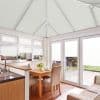 White pleated blinds cover the conservatory roof, windows, and French doors, drawn closed and diffusing daylight over a bright dining area with wooden table, upholstered chairs, sink, and garden beyond.