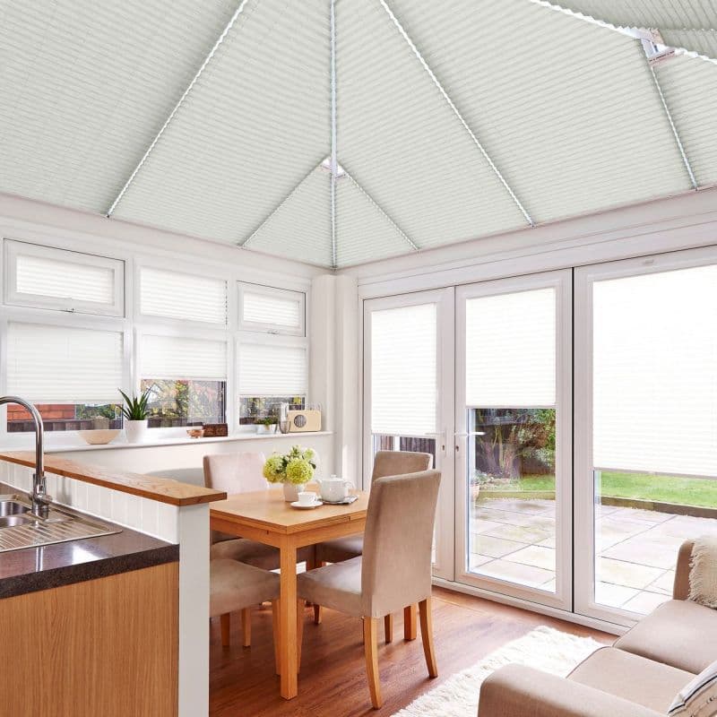 White pleated blinds cover the conservatory roof, windows, and French doors, drawn closed and diffusing daylight over a bright dining area with wooden table, upholstered chairs, sink, and garden beyond.