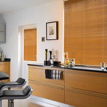 Wooden horizontal Venetian blinds cover a large window and small door window at right, closed and filtering warm daylight. Modern kitchen with light-wood cabinets, black countertop, bar stools.