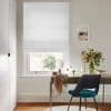 White linen Roman blind, fully lowered with subtle folds, filtering soft daylight through the window; compact home office with wooden desk, teal chair, a vase of flowers, and framed seaside print.