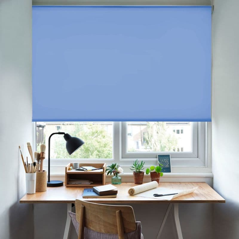 A blue roller blind lowered across a double window, opaque fabric filtering daylight over a wooden desk with lamp, potted plants, art supplies and chair in a tidy study.
