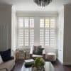 White plantation shutters cover a three-section bay window—two-tier panels with horizontal louvers—partly closed, upper slats slightly open, filtering soft daylight into a neutral living room with an armchair and ottoman.