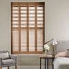 Wooden plantation shutters—four-panel, louvered—cover a tall window, slats partially open and filtering light; bedroom corner with upholstered bed, bedside table, lamp, and gray armchair.