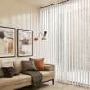 Sheer vertical blinds drawn across a floor-to-ceiling corner window, softly filtering daylight; beige living room with mid-century swing wall lamp, abstract framed art, neutral sofa with textured cushions and throw.