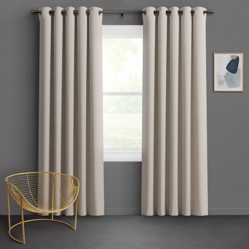 Cream grommet-top curtains, floor-length and pleated on a metal rod, parted to reveal bright window; hanging and filtering diffused daylight in a gray-walled minimalist room with gold wire chair.