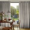 Pleated grey patterned curtains, parted at center, framing and admitting soft daylight; wooden dining table set for breakfast beneath a pendant lamp, chairs facing a sunlit garden.