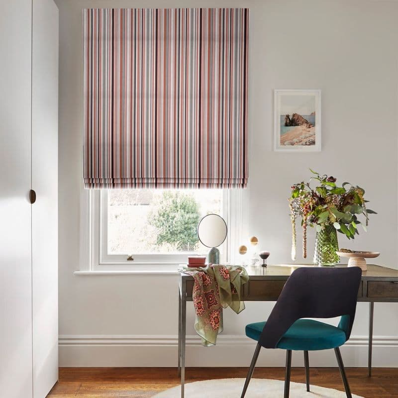 A striped Roman blind, lowered over the window with red, navy and cream vertical bands, filtering soft daylight; a home office desk with teal chair and vase of flowers.