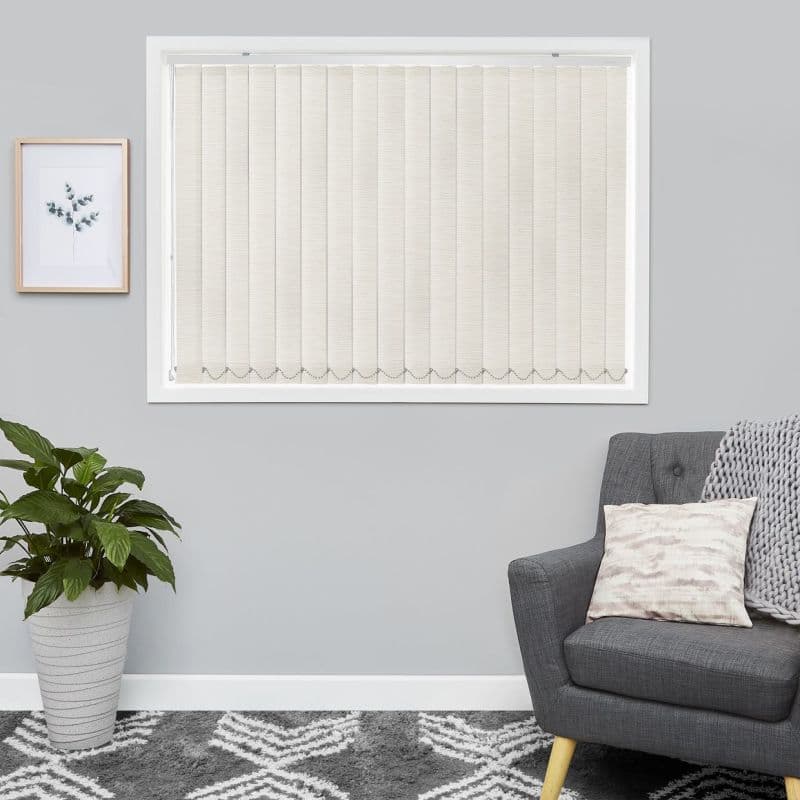 Beige vertical blinds covering the framed window, fully closed with scalloped bottom chain, softly filtering daylight; in a grey living room with a potted plant, botanical print, armchair, rug.