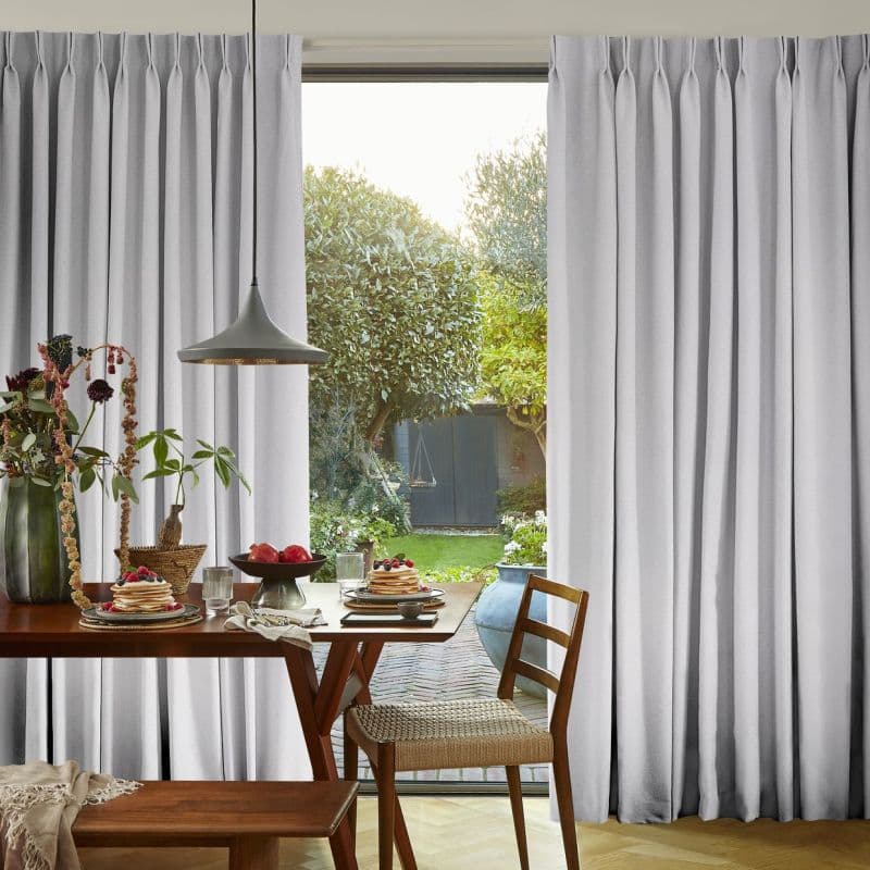 Floor-length pleated gray curtains, pulled aside at center to frame a glass door, admitting soft daylight. Dining table with pendant lamp, pancakes on plates, wooden chairs, leafy backyard view.