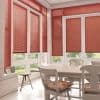 Red horizontal slatted blinds covering tall bay windows and doors, mostly lowered and filtering daylight; inside, a bright dining nook with white table, chairs, tiled floor and glimpses of garden.