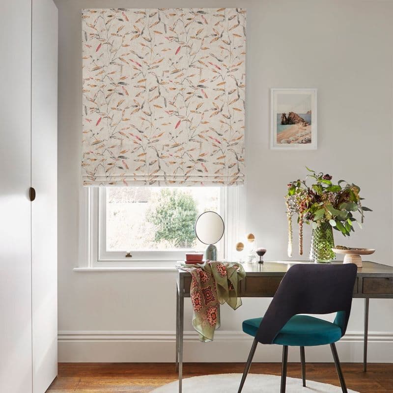 Patterned Roman blind, fully lowered, pale fabric with delicate leaf motif, filtering soft daylight above a desk with mirror, vase of flowers and teal chair in a bright neutral office.