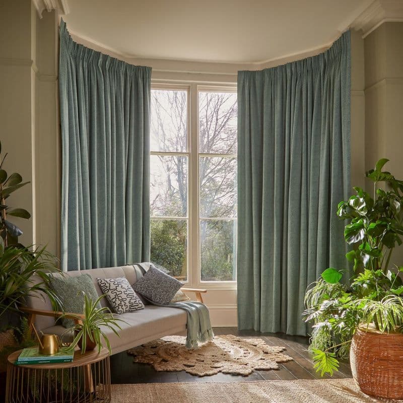 Full-length pleated teal curtains framing a tall bay window, hung on a track and drawn partly aside, filtering daylight into a cozy living room with sofa, rug and houseplants.