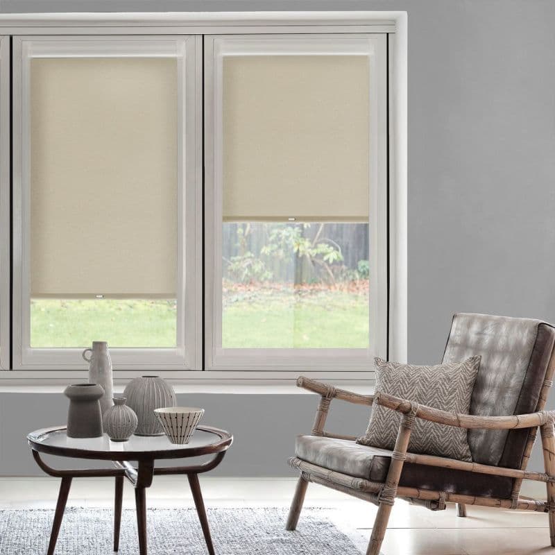 Beige roller blinds, both lowered (left fully, right partially), filtering daylight through two tall windows; muted living room with round wooden table and ceramics, rattan-leather armchair and grassy yard beyond.