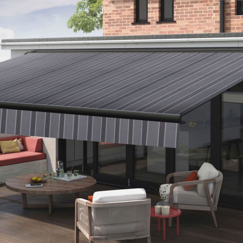 Retractable striped fabric awning, dark grey with diagonal pattern and striped valance, fully extended over sliding glass doors—shading a sunlit modern patio with wooden deck, cushioned seating and coffee table.