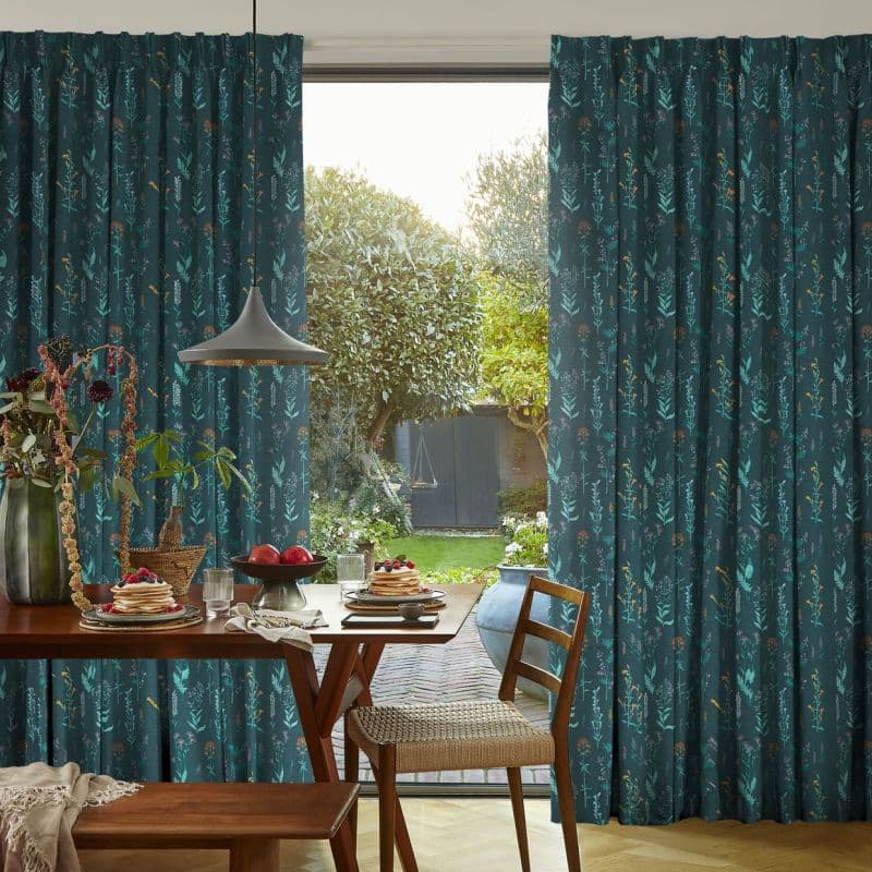 Dark teal, patterned floor-to-ceiling curtains, drawn aside to frame a sliding glass door; allowing daylight into a dining area with wooden table set for breakfast, pendant lamp, and garden view.