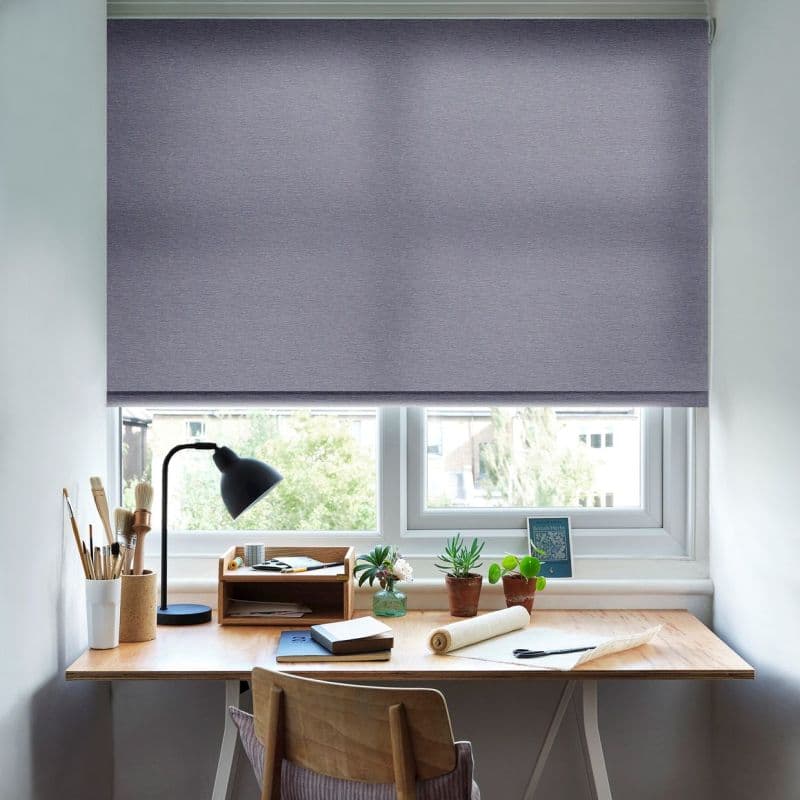 Grey textured roller blind, lowered covering most of the window, softly diffusing daylight; over a tidy wooden desk with lamp, brushes, notebooks, potted plants and a suburban view.