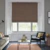 Patterned fabric Roman blind (grey with orange lattice) lowered over bay window, partially filtering bright daylight; living room with grey walls, sofa, black armchair, coffee table and cushions.