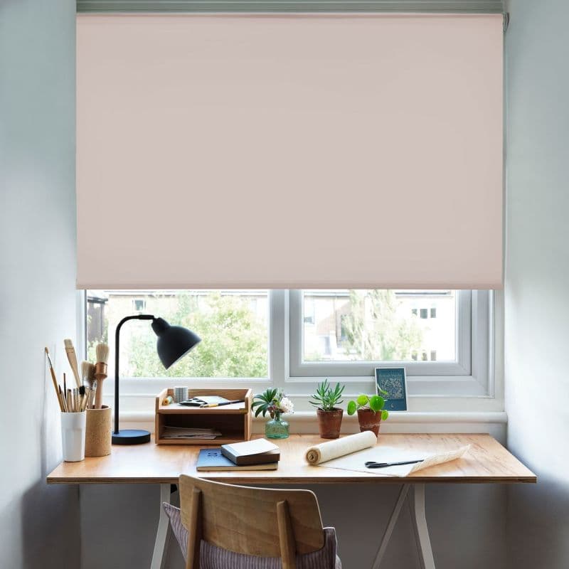 Pale blush roller blind, fully lowered across a double‑pane window with smooth fabric, blocking and softly diffusing daylight; a small home desk beneath holds a lamp, plants, and brushes.