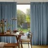 Full-length pleated blue curtains, parted slightly, letting soft daylight through a sliding glass door; a wooden dining table set with pancakes, a pendant lamp, and a lush garden view.