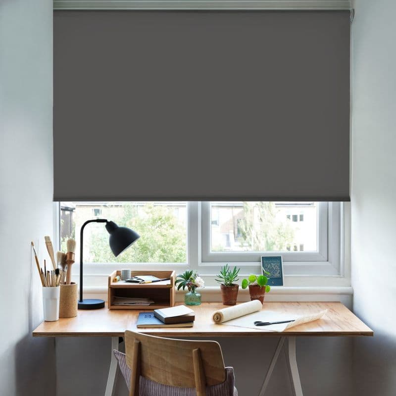 Grey roller blind lowered to just above the sill, opaque and blocking light; above a wooden desk with lamp, potted plants, art brushes and notebooks in a lit home workspace.