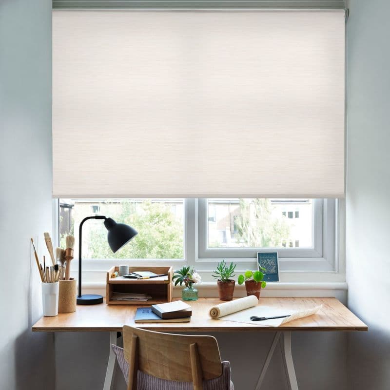 A cream roller blind, almost fully lowered and lightly textured, diffusing daylight—covering a double window—above a small wooden desk with a lamp, art brushes, notebooks and potted succulents, soft suburban view.