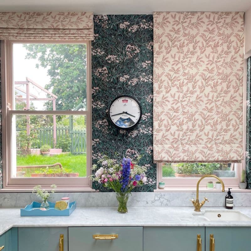 Pair of patterned Roman blinds—left raised, right partially lowered—soft pink botanical fabric filtering daylight over a kitchen sink with marble counter, floral wallpaper and visible green garden outside.