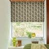 Roman blind with brown‑beige geometric chain pattern, lowered to mid-window, partially filtering daylight; bedroom sill holds green glass vases, potted plant and small fan, with a leafy garden beyond.