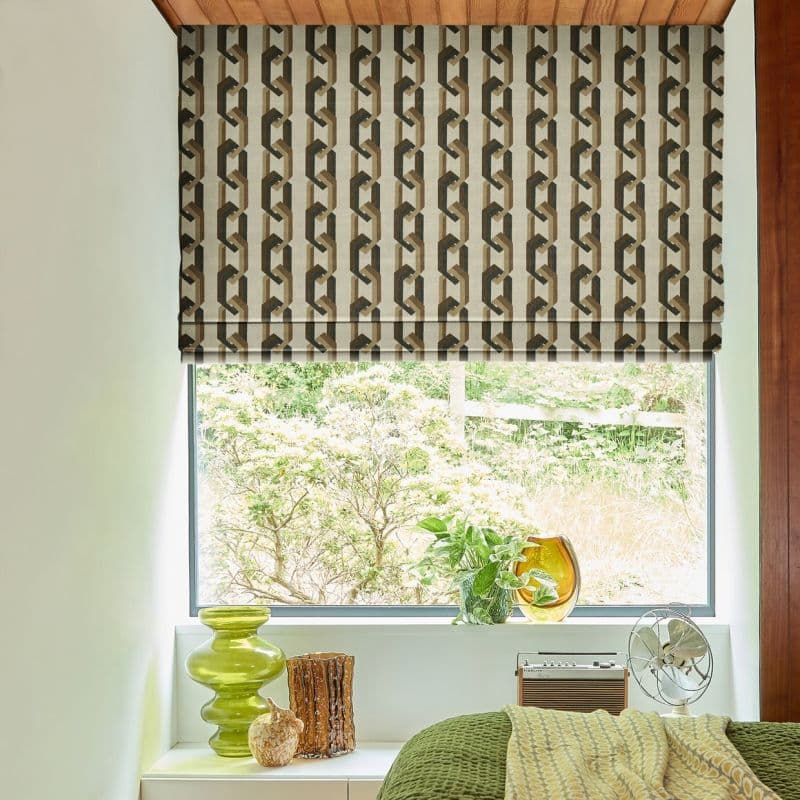 Roman blind with brown‑beige geometric chain pattern, lowered to mid-window, partially filtering daylight; bedroom sill holds green glass vases, potted plant and small fan, with a leafy garden beyond.