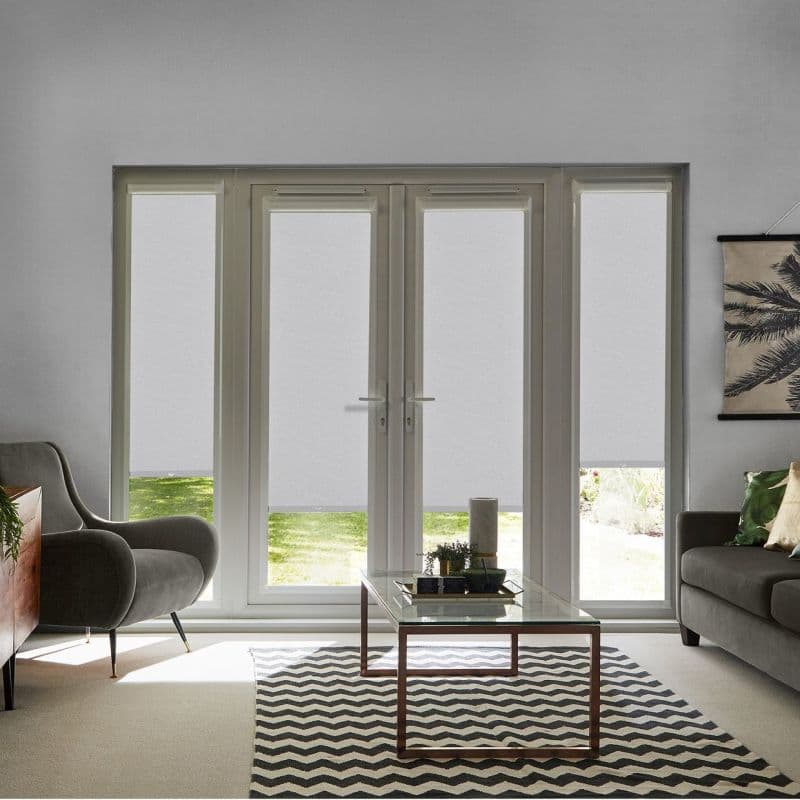 Light-grey roller blinds lowered across four tall French doors and side panels, leaving narrow uncovered bottoms; filtering daylight into a living room with sofas, coffee table, chevron rug, garden view.