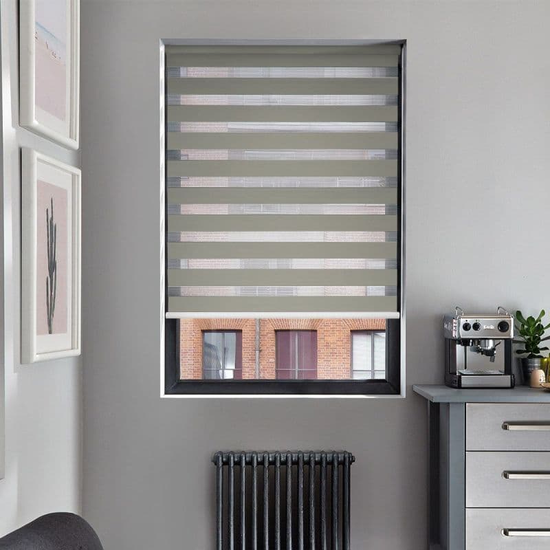 Zebra roller blind with alternating opaque gray and sheer horizontal bands, mostly lowered and filtering light over a window in a gray kitchen nook with radiator and espresso machine.