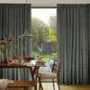 Floor-to-ceiling, patterned pleated curtains, dark blue chevron, partially drawn to frame a sliding glass door; filtering morning light over a dining table set with pancakes, pendant lamp, and backyard garden.