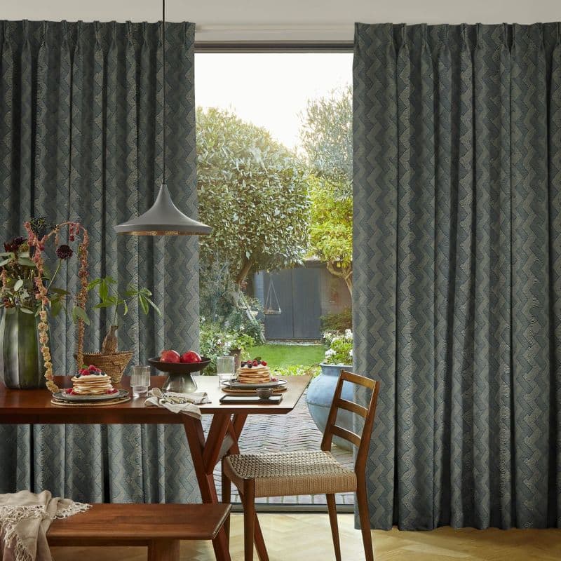Floor-to-ceiling, patterned pleated curtains, dark blue chevron, partially drawn to frame a sliding glass door; filtering morning light over a dining table set with pancakes, pendant lamp, and backyard garden.