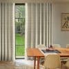 Patterned beige curtains, mostly drawn with a narrow central opening filtering daylight; covering a tall multi‑pane window overlooking a green yard, in a dining room with wooden table and chairs.