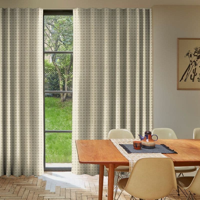 Patterned beige curtains, mostly drawn with a narrow central opening filtering daylight; covering a tall multi‑pane window overlooking a green yard, in a dining room with wooden table and chairs.