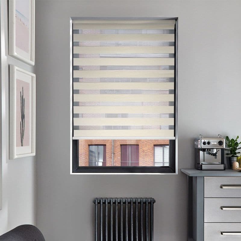 Beige-and-gray zebra roller blind, fully lowered with alternating opaque and sheer horizontal stripes, filtering daylight; modern gray-kitchen corner visible — countertop with espresso machine and plant, radiator below, brick-building view.