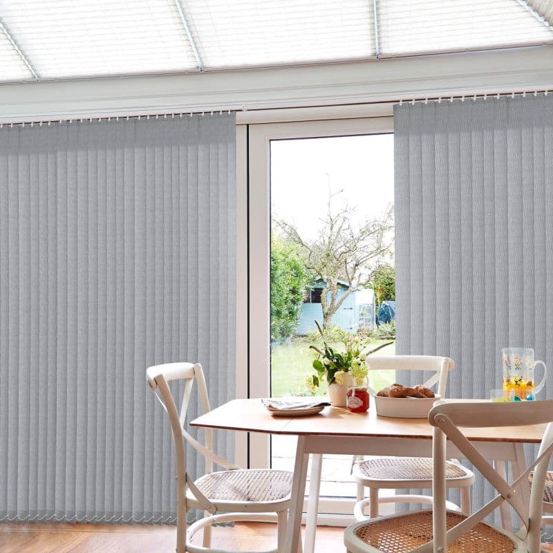 Grey fabric vertical blinds, floor-to-ceiling and rail-mounted, partially drawn across sliding glass doors, filtering daylight; small dining table and chairs in foreground, garden and shed visible through central gap.