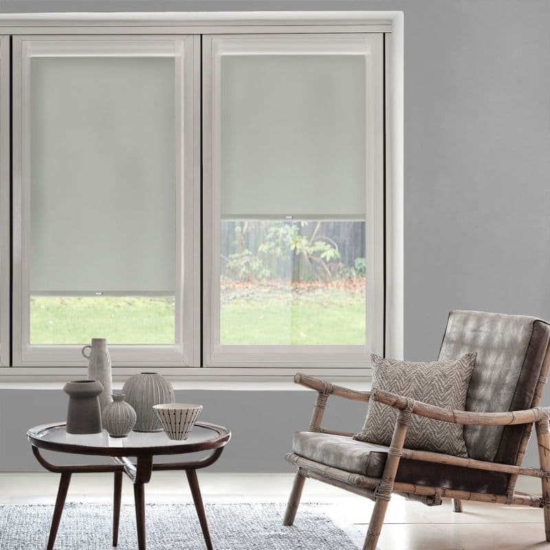 Roller blinds covering a double-pane window, lowered halfway with light-filtering white fabric; living room with neutral decor, wooden side table, ceramic vases, leather armchair, soft daylight and grassy backyard view.