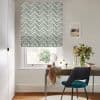 Chevron-patterned Roman blind, lowered to mid-window, mosaic teal, gold and black chevrons; filtering soft daylight over a wooden desk with teal chair, vase of flowers and a framed seaside print.