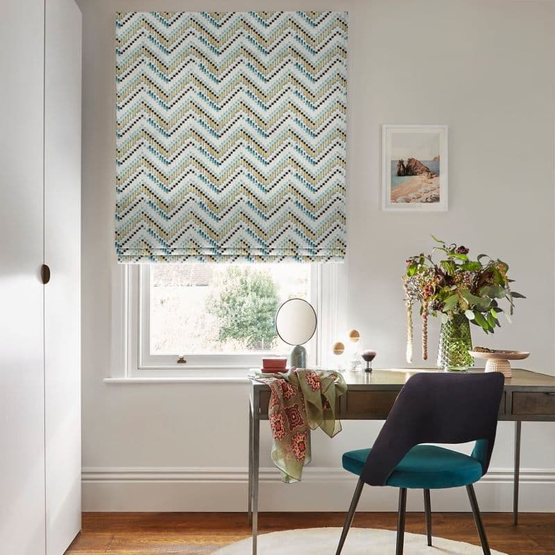 Chevron-patterned Roman blind, lowered to mid-window, mosaic teal, gold and black chevrons; filtering soft daylight over a wooden desk with teal chair, vase of flowers and a framed seaside print.