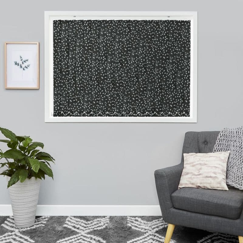 Dark vertical blinds with pale leaf print, fully closed and blocking light; in a gray living room with a gray armchair, chunky throw, potted plant and framed botanical print.