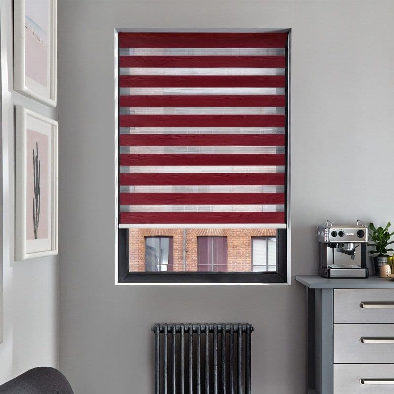 A burgundy-and-sheer striped zebra roller blind, lowered across the window with alternating horizontal bands, filters light and partly obscures a brick-building view in a gray kitchen corner with espresso machine.