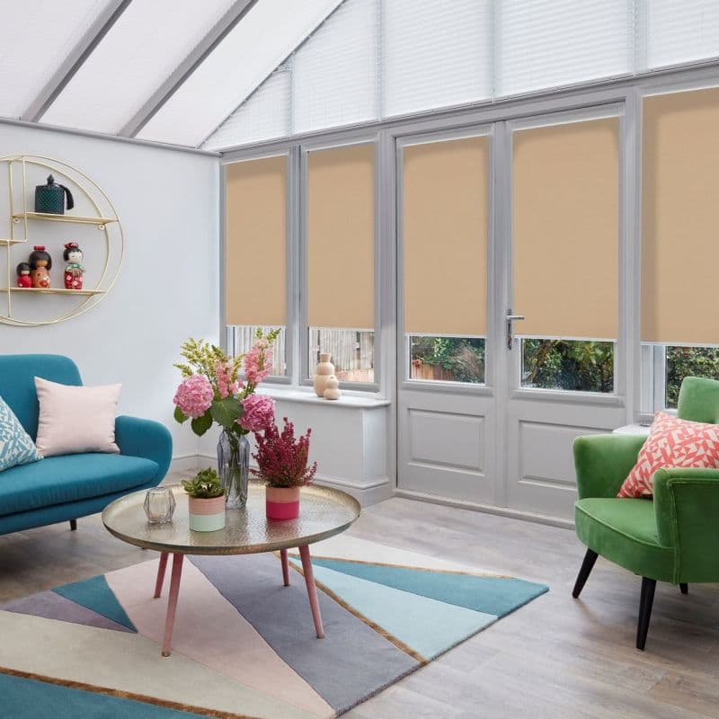 Beige roller blinds on tall French doors and windows, pulled down to mid-height with white pleated blinds above; blocking direct sun and softening light in a bright conservatory living room.