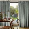 Striped pleated curtains, blue-gray and cream stripes, hang either side of a tall glass door, partially open, filtering morning light onto a dining table set for breakfast; sunlit garden beyond.